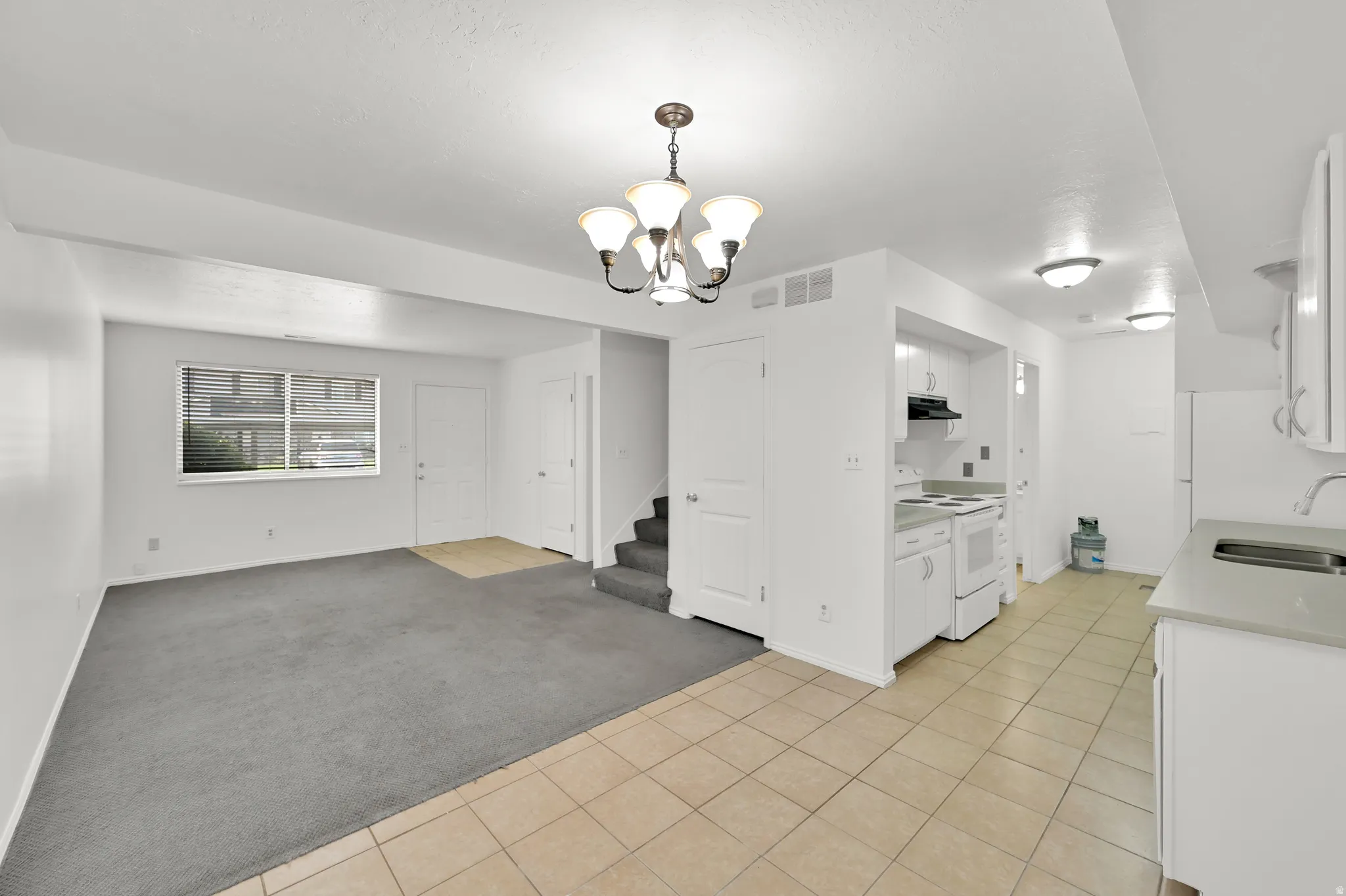 Bright open-concept living area with a contemporary chandelier, large window with blinds, and durable tile flooring in the kitchen