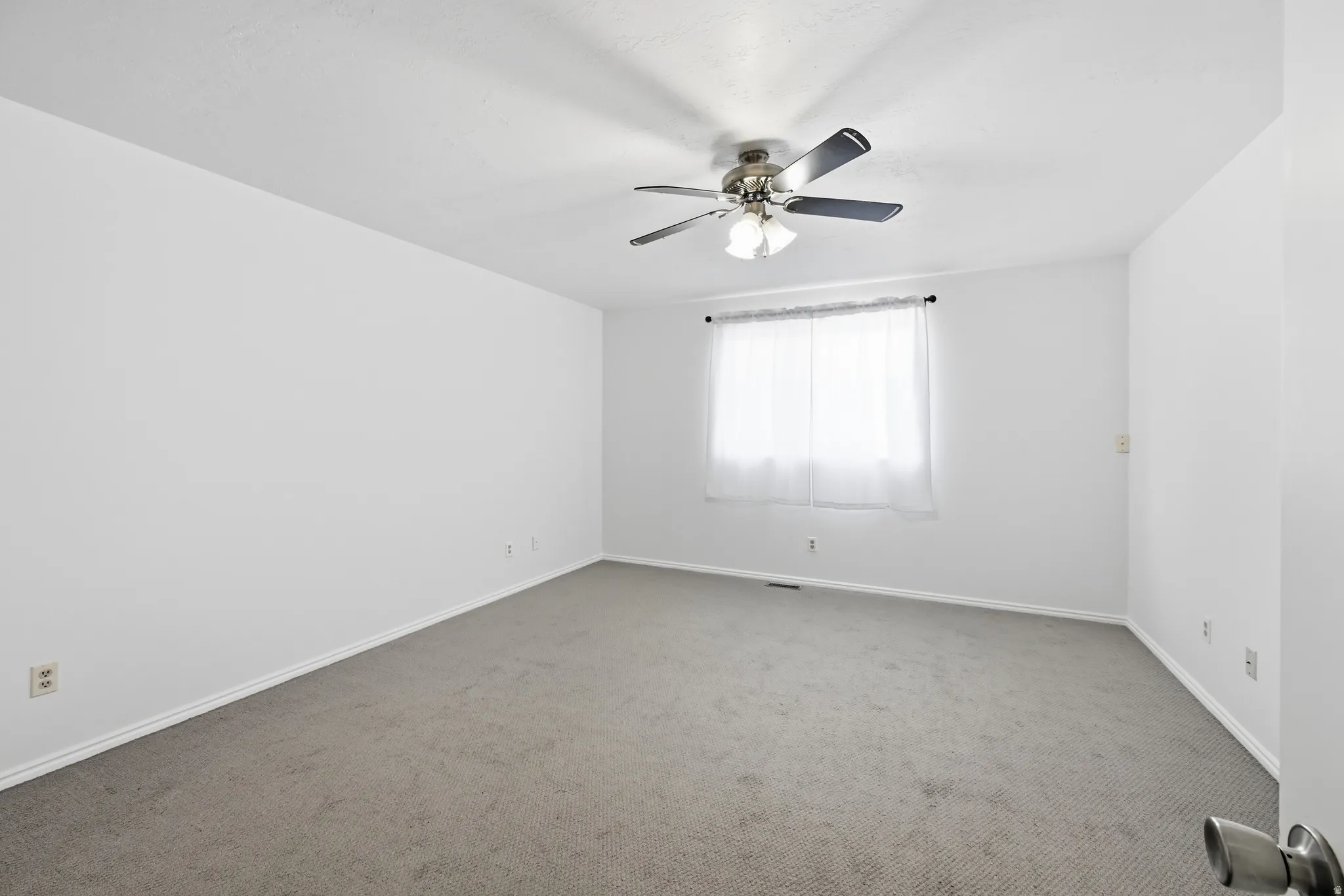 Neutral wall paint, ceiling fan with integrated lighting, window with light-filtering curtains, and grey carpeting