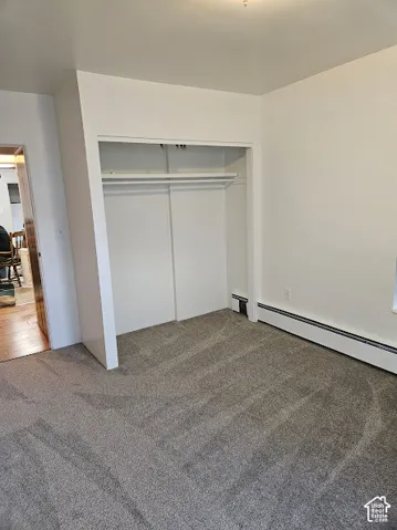Plush grey carpeting, white painted walls, built-in closet with shelving and hanging rod, and baseboard heating