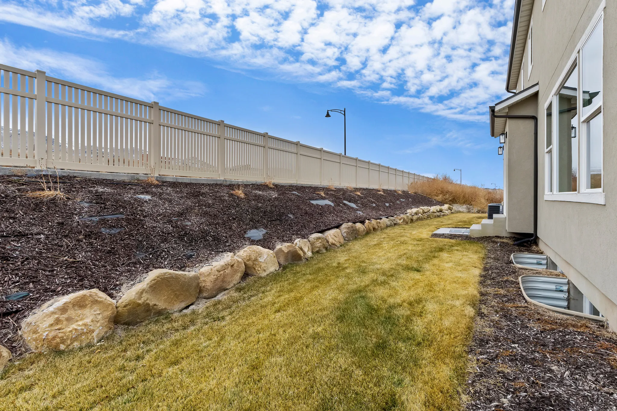 Rear yard with sloped landscape, rock retaining wall, privacy fence, and multiple window wells