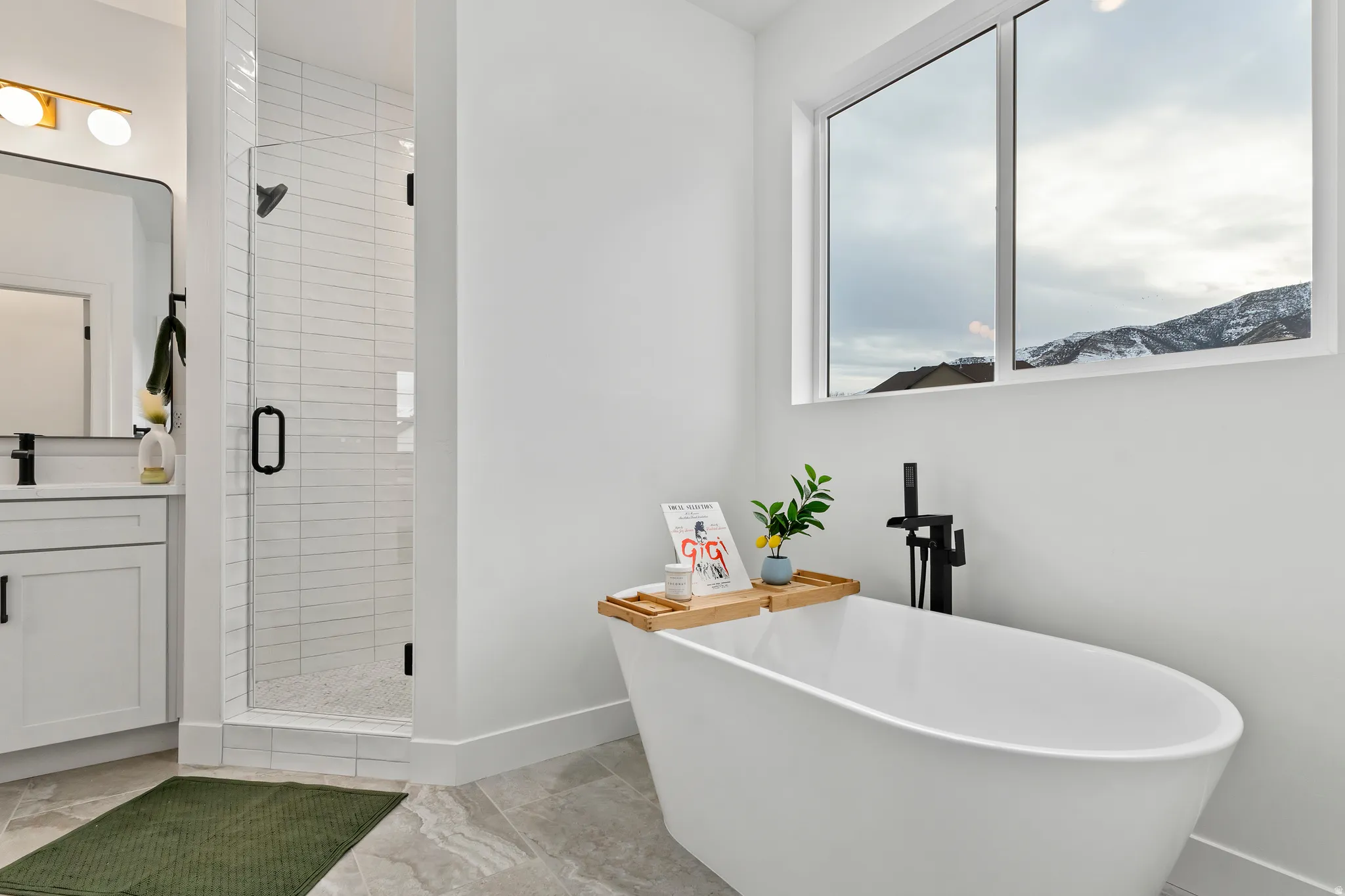 Freestanding soaking tub with a floor-mounted matte black faucet, glass-enclosed shower with subway tile, white vanity with matte black hardware, and large window