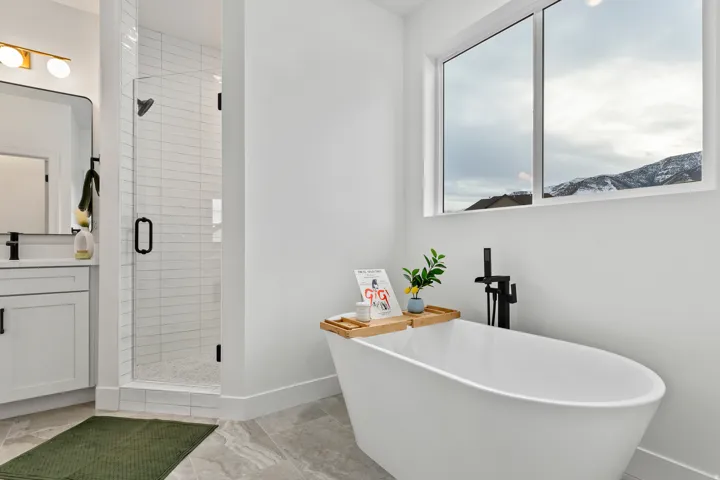Freestanding soaking tub with a floor-mounted matte black faucet, glass-enclosed shower with subway tile, white vanity with matte black hardware, and large window