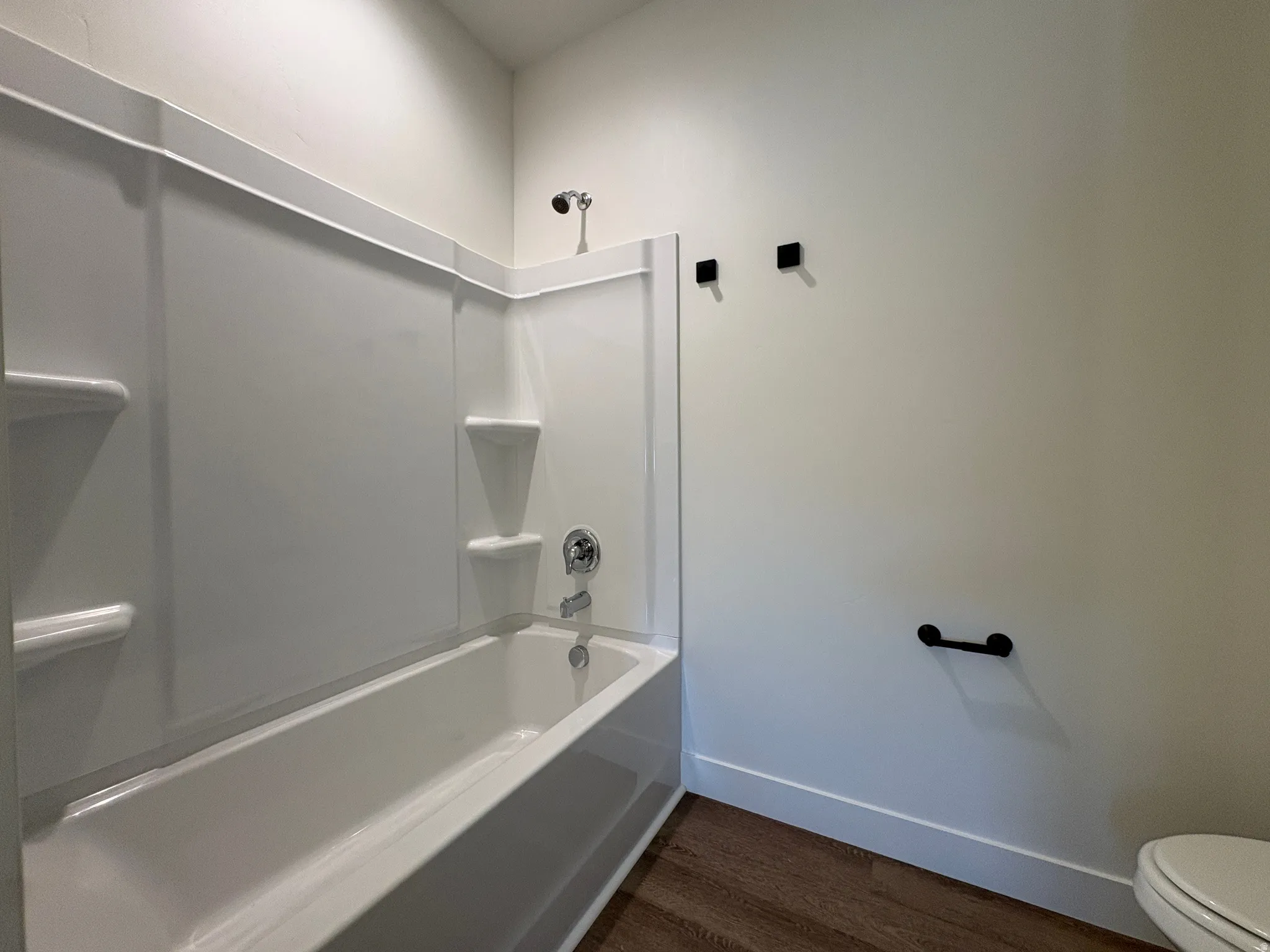 White tub-shower combination with integrated shelving, chrome fixtures, and wood-finish flooring