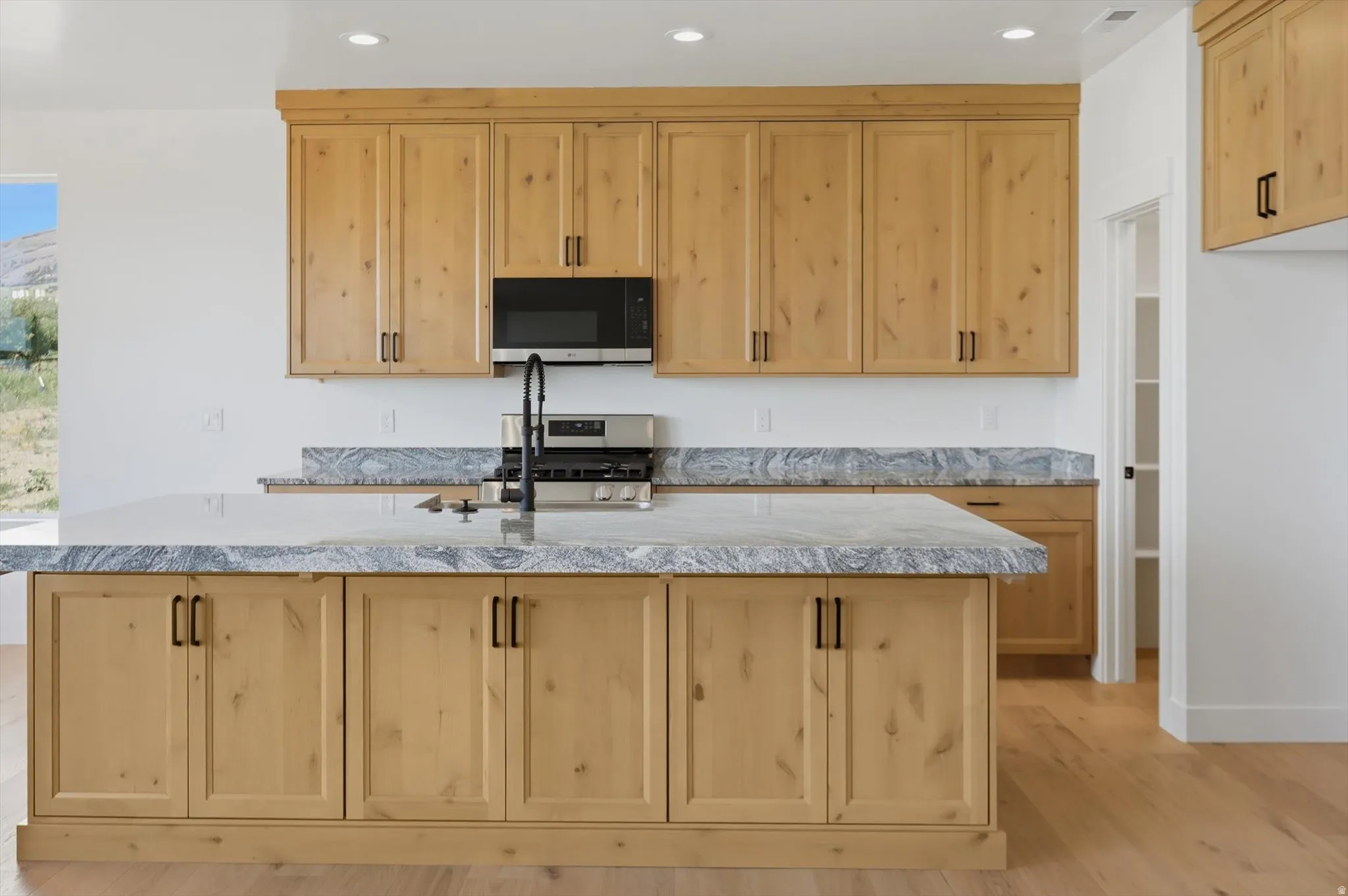 Natural wood-finish cabinetry with black hardware, stone countertops with matching backsplash, stainless steel range and microwave, and wood-finish flooring