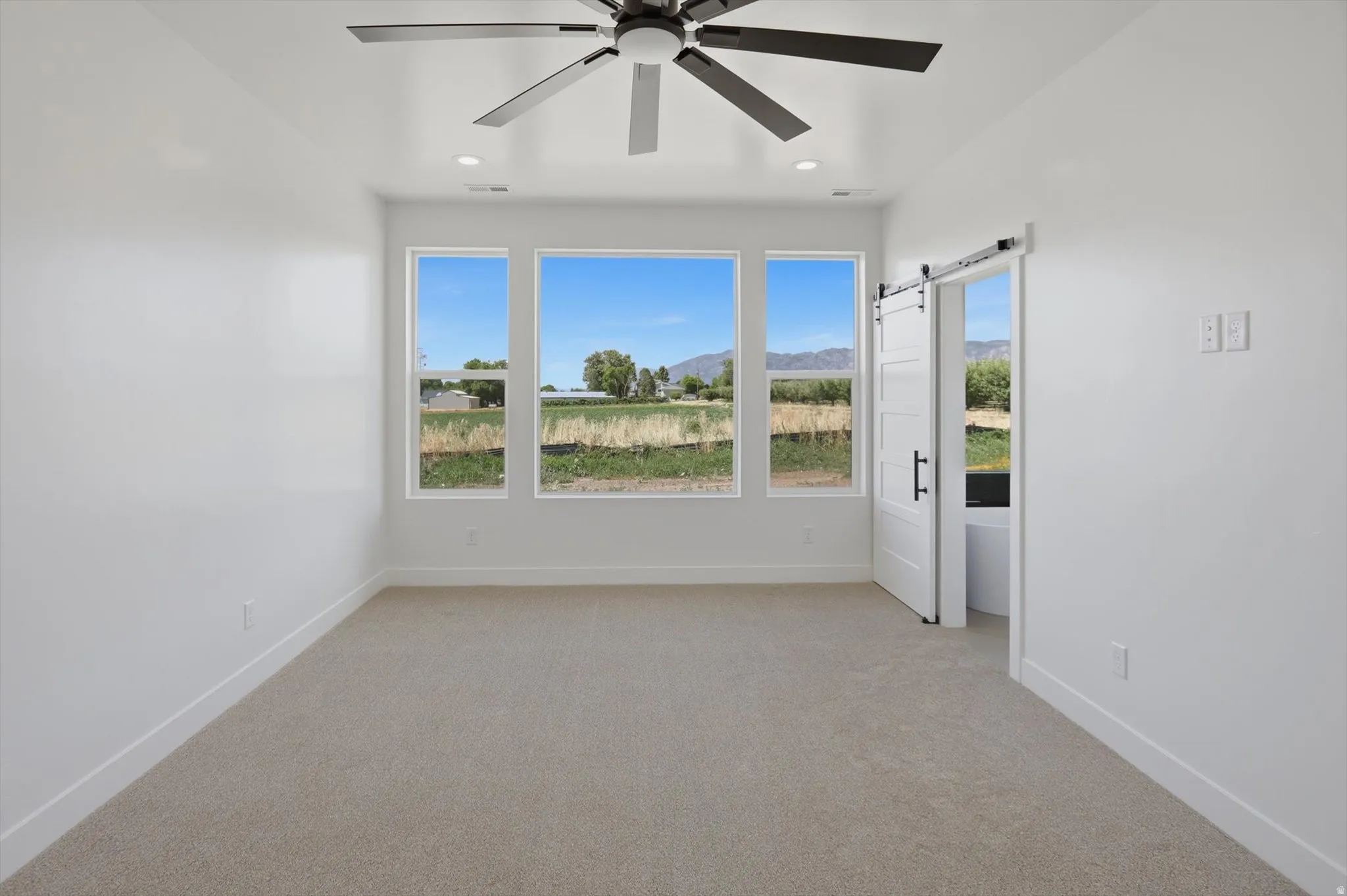 Recessed lighting, ceiling fan with dark finish, three large windows, carpeted flooring, and a barn door with dark hardware