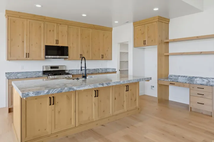 Light wood cabinetry throughout, featuring a large kitchen island with a dark matte faucet, integrated stainless steel appliances, and a built-in desk area with open shelving