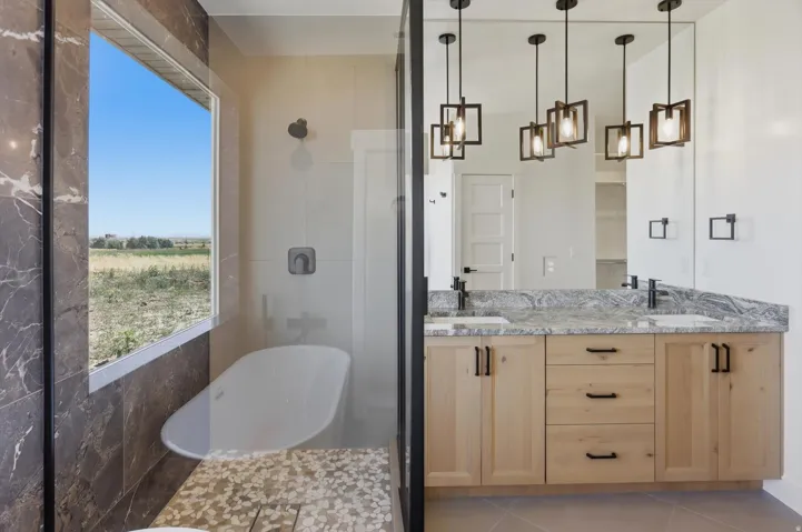 Freestanding soaking tub with a large picture window, a walk-in shower with stone tile, a dual vanity with light wood cabinetry, and pendant lighting fixtures