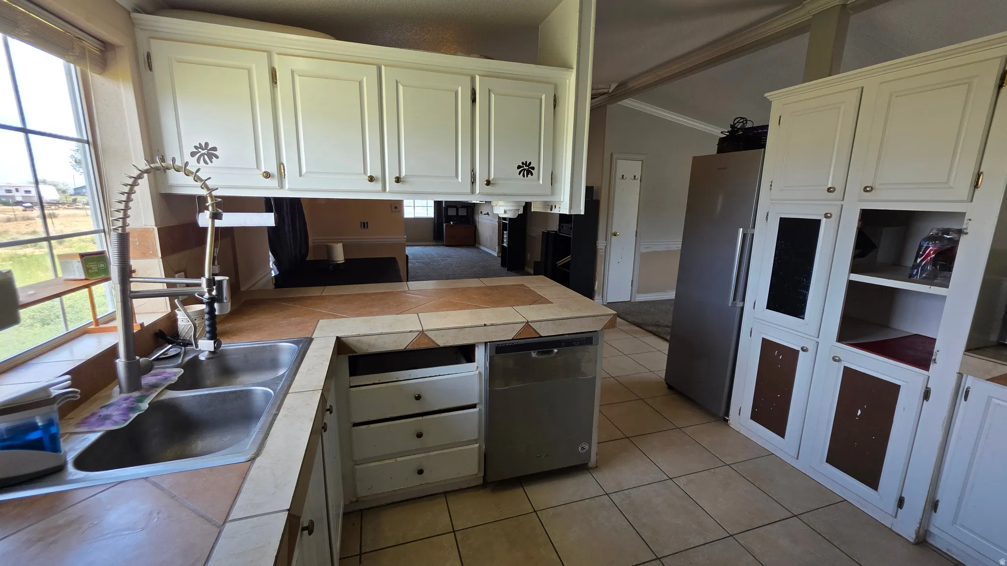 White cabinetry with gold-tone hardware, tile countertops, dual basin stainless steel sink with a high-arch faucet, integrated dishwasher, and stainless steel refrigerator