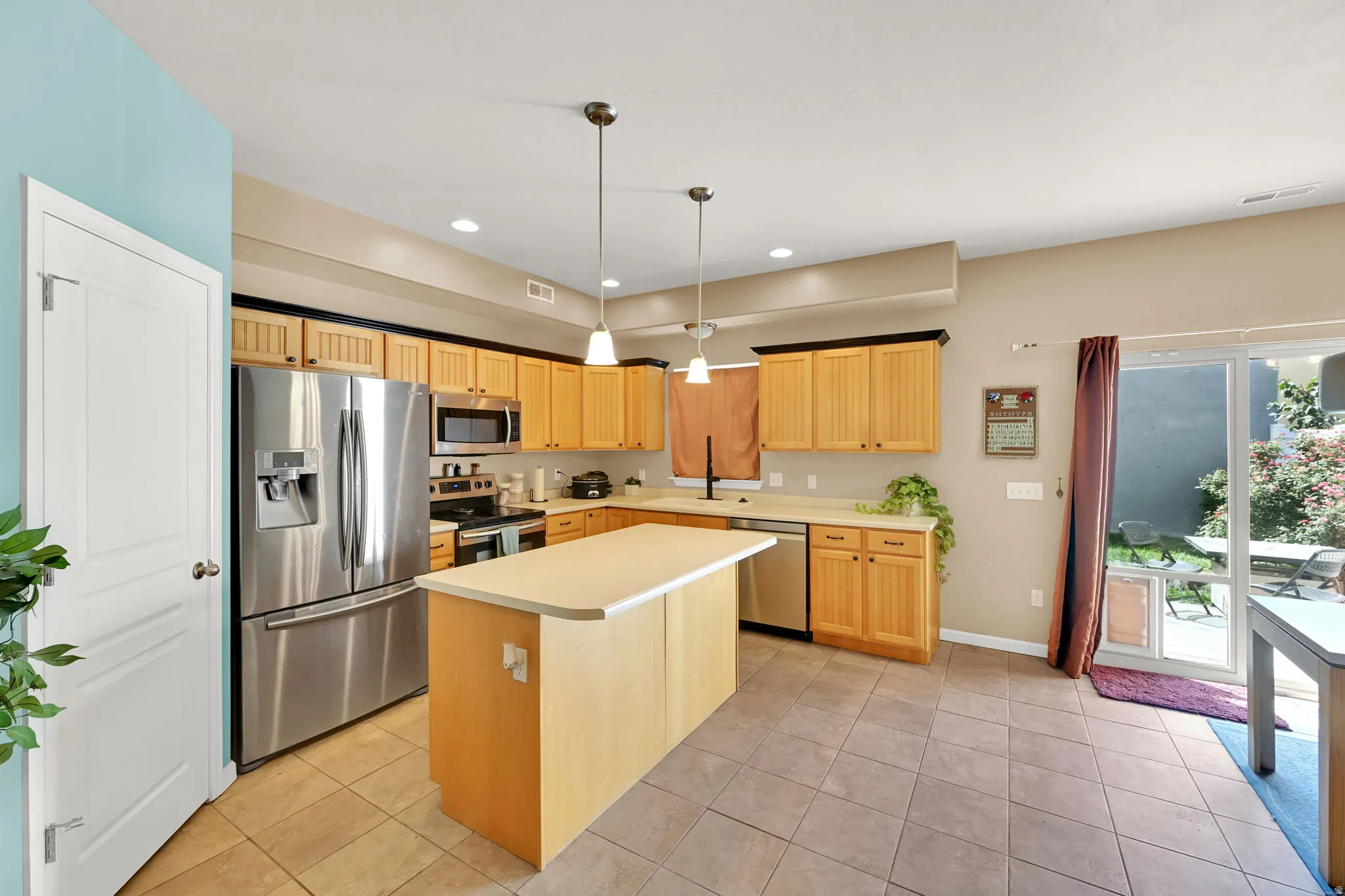 Kitchen featuring light wood cabinetry, stainless steel appliances, a center island with countertop, tile flooring, and pendant lighting