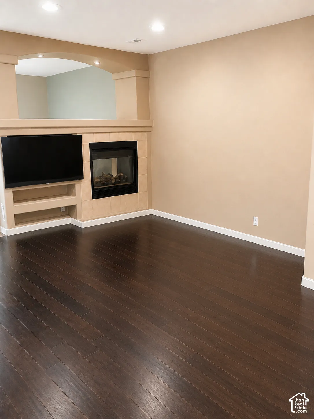 Dark wood-finish flooring, built-in media center with shelving, integrated fireplace, recessed lighting, and an arched architectural opening