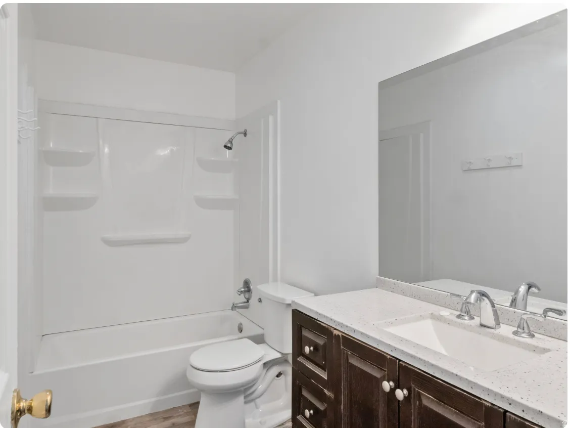 White acrylic tub and shower combination with built-in shelving, dark wood-finish vanity with white knobs, white rectangular sink, and white speckled countertop