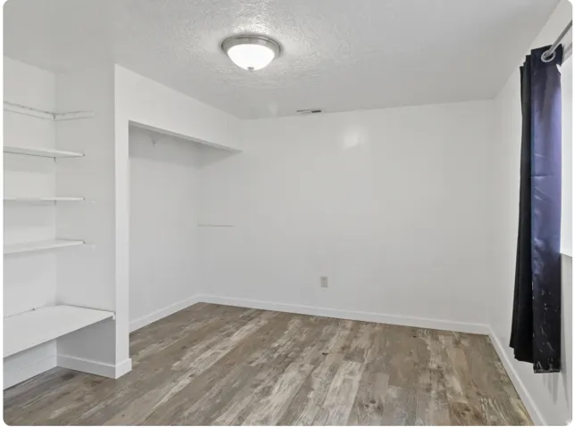 White walls with wood-finish flooring, featuring built-in shelving, a recessed clothing rod, and a ceiling-mounted light fixture