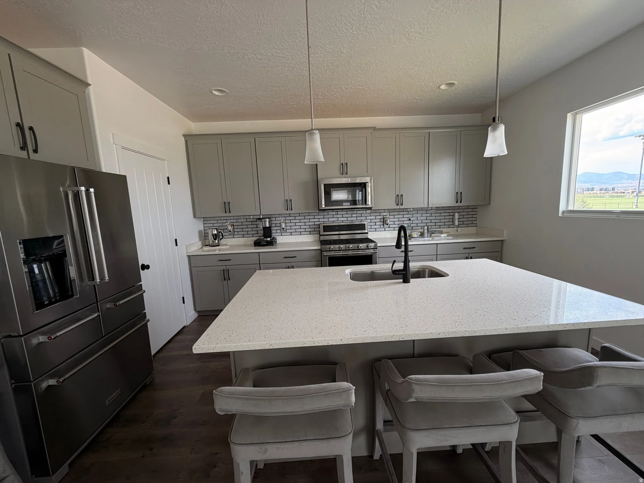 Stainless steel appliances, grey cabinetry, mosaic tile backsplash, central island with integrated sink, and wood-finish flooring
