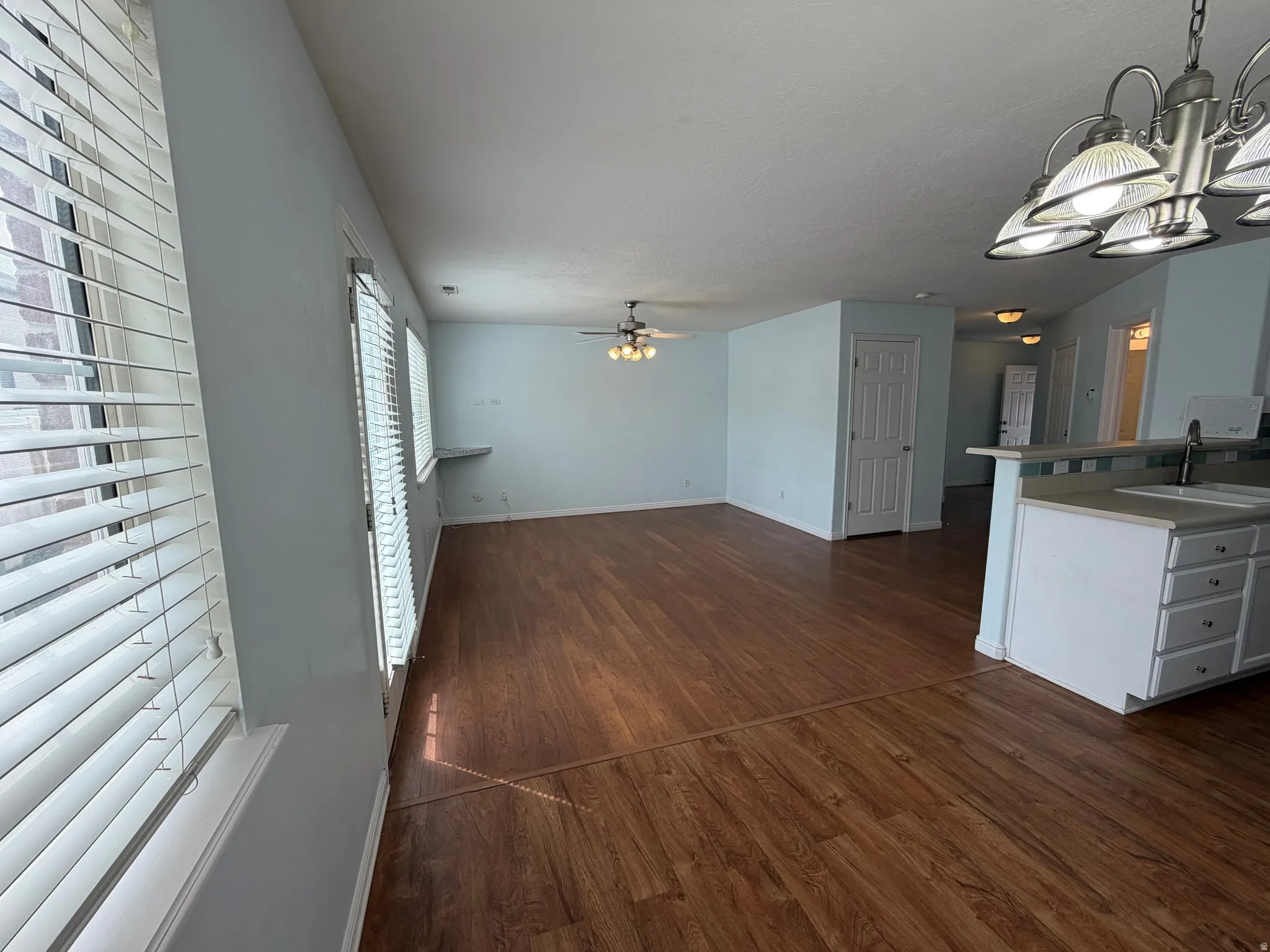 Open-concept living area featuring wood-finish flooring, multiple windows with blinds, a ceiling fan, and a kitchen peninsula with a sink and overhead lighting
