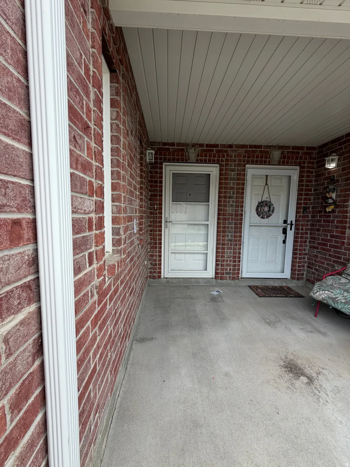 Red brick exterior wall, concrete flooring, white soffit ceiling, and two entry doors with storm doors