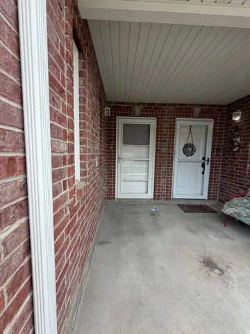 Red brick exterior wall, concrete flooring, white soffit ceiling, and two entry doors with storm doors