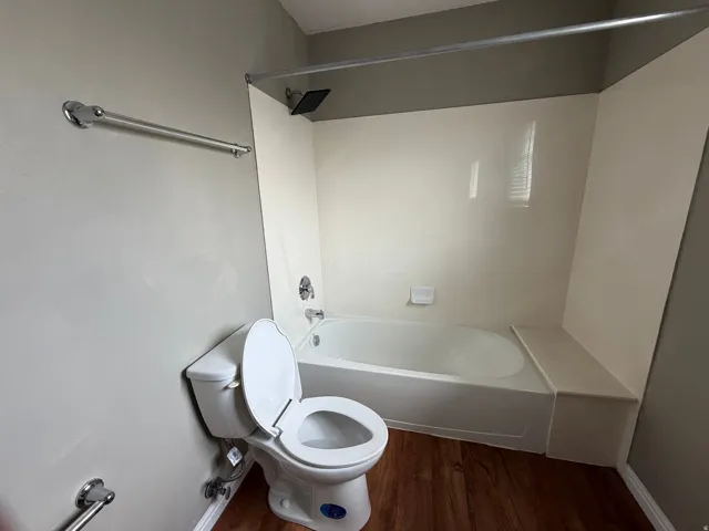 Tub and shower combination with a black shower head, white tiled surround, and wood-finish flooring