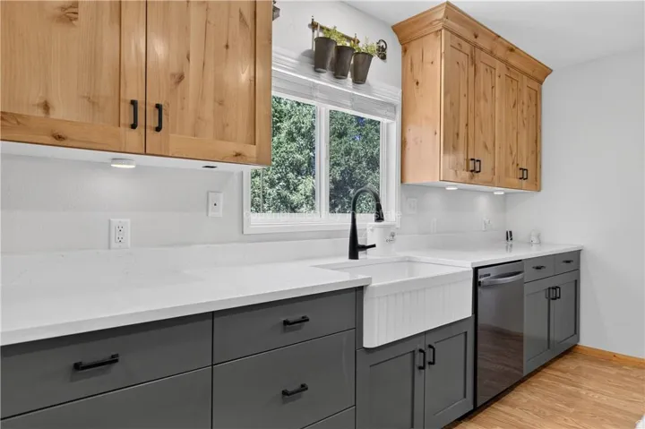 Two-tone cabinetry with light wood upper cabinets and dark gray lower cabinets, white countertops, white apron-front sink, black gooseneck faucet, and stainless steel dishwasher