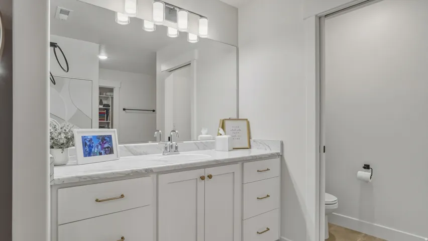 Double vanity with a white countertop and integrated sink, under-mount cabinetry with gold-tone hardware, and a wall-mounted light fixture above a large mirror