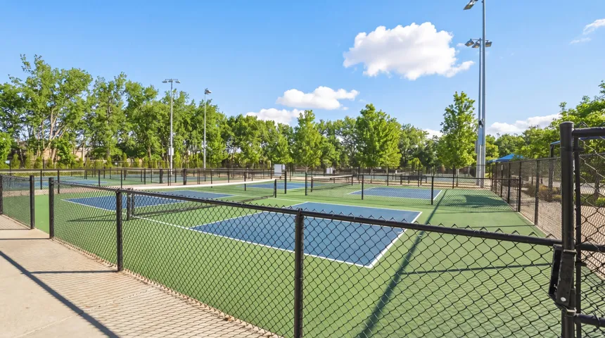 Multiple outdoor courts with chain-link fencing, featuring green and blue playing surfaces, perimeter lighting, and mature trees