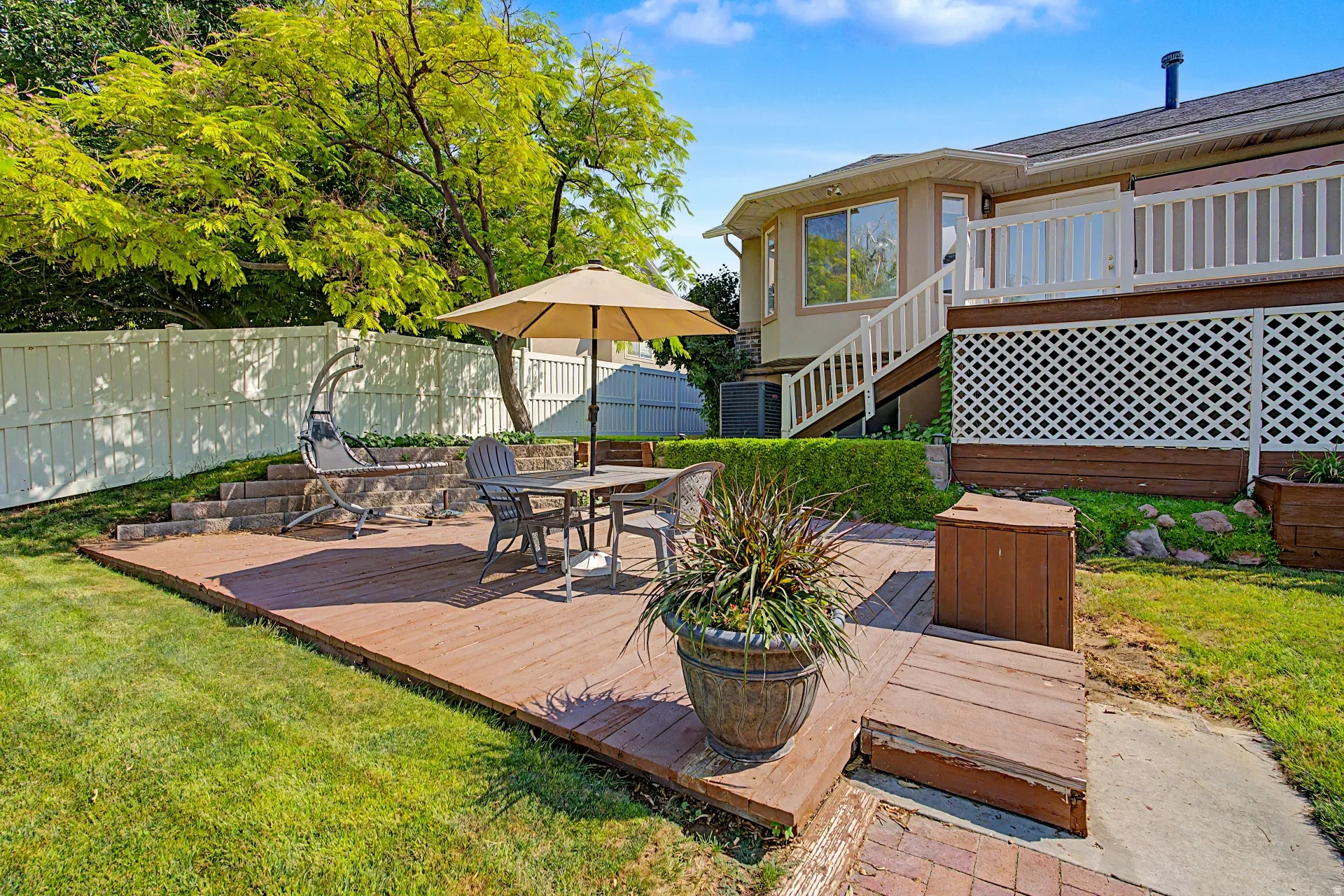 Expansive backyard featuring a wood-finish deck, white privacy fencing, mature trees, and a tiered landscaped area