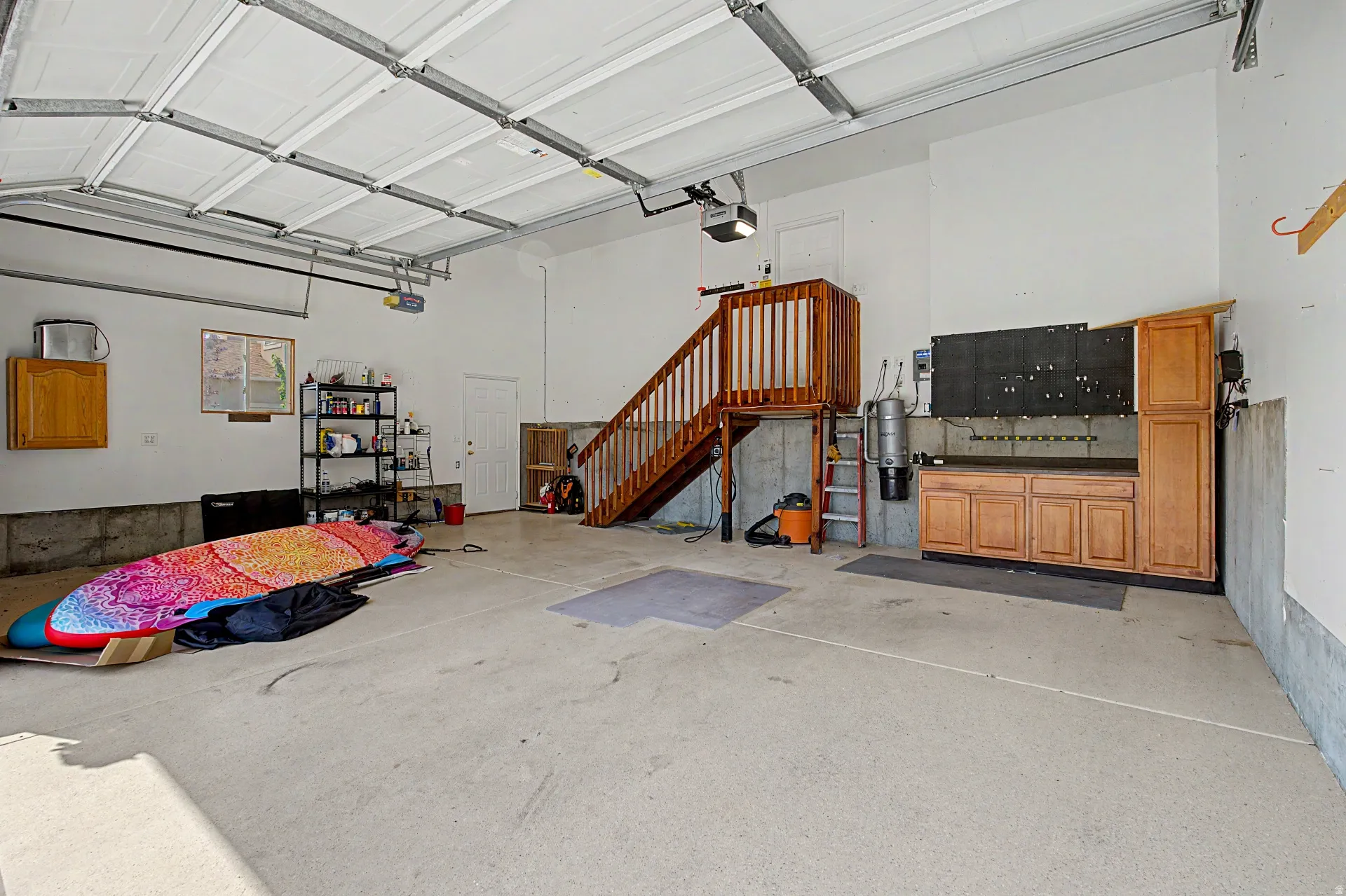 Spacious garage with polished concrete flooring, overhead sectional garage door with automatic opener, wood cabinetry storage, wooden stairs with railing providing attic access, and bright overhead lighting from recessed fixtures and skylights