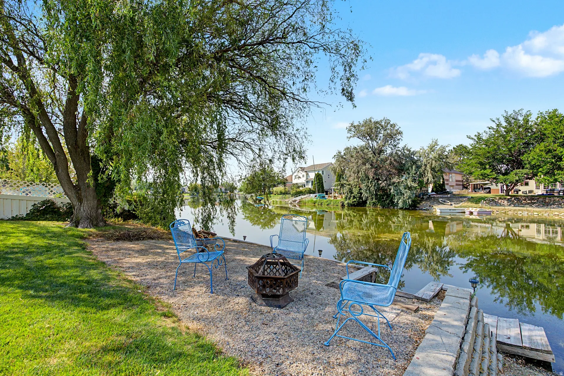 Expansive waterfront property featuring a manicured lawn, established weeping willow, gravel patio, and stone retaining wall
