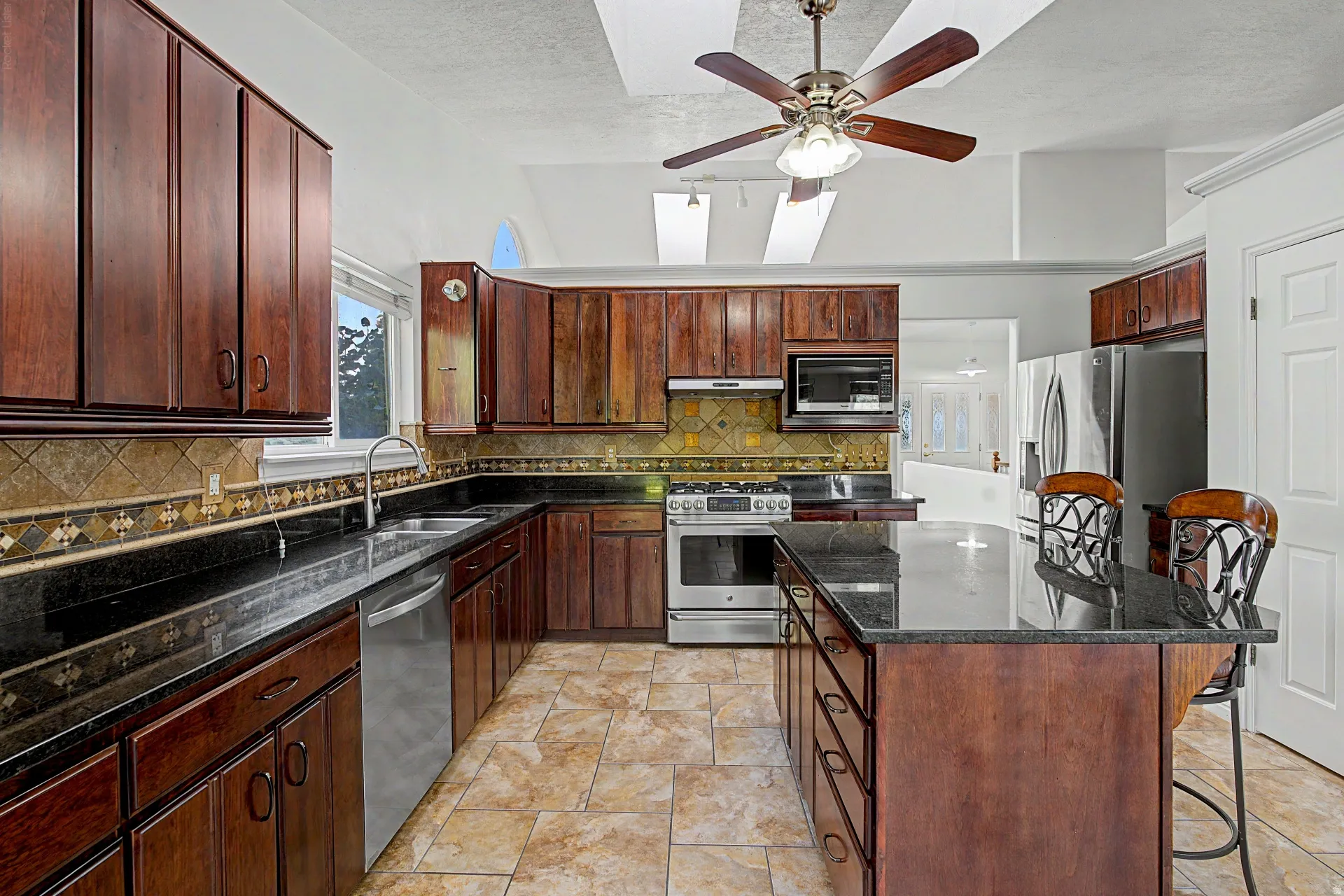 Kitchen featuring dark wood cabinetry, black countertops, stainless steel appliances, a tiled backsplash, and a ceiling fan