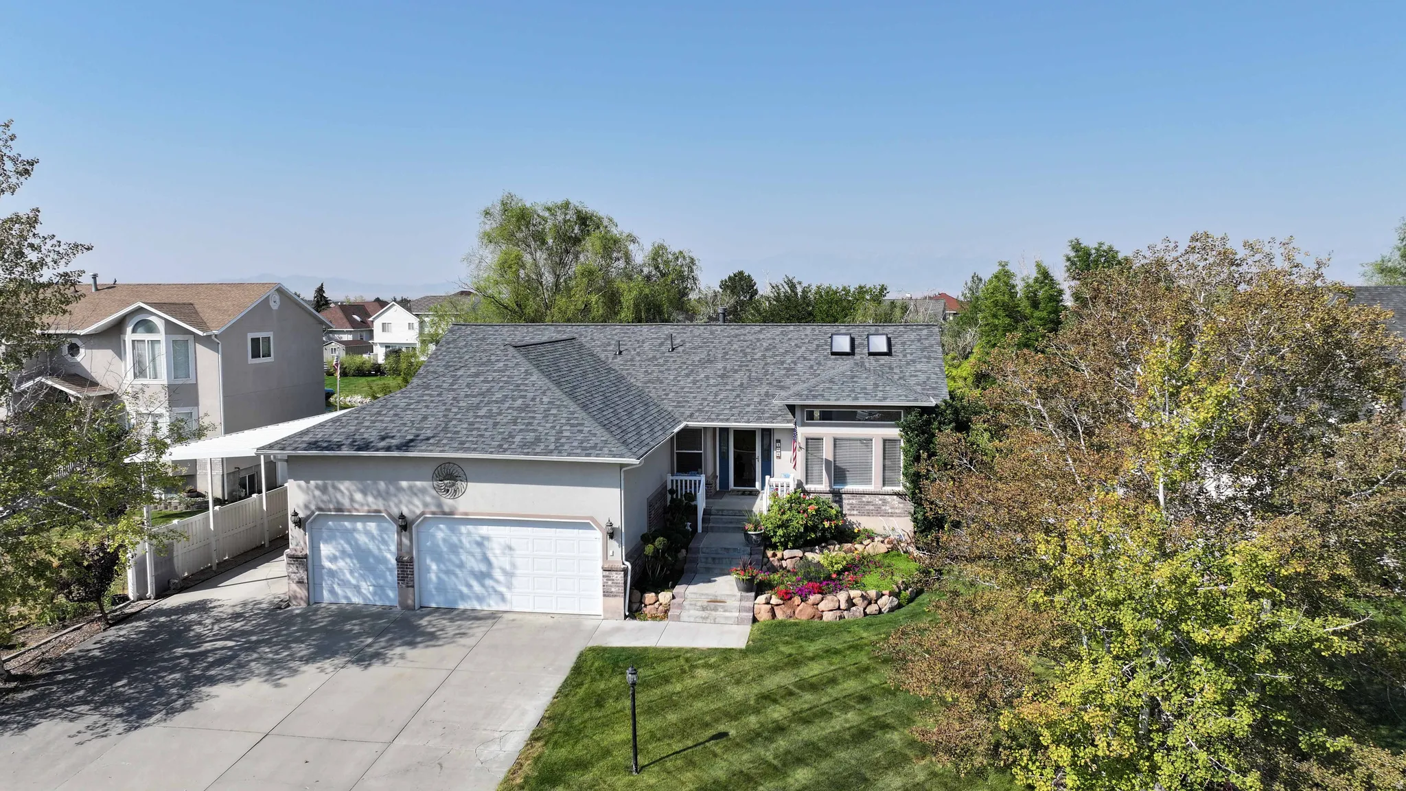 Two-car garage with white paneled doors, concrete driveway, stone facade accents, front yard landscaping with mature plantings, and a pitched shingle roof