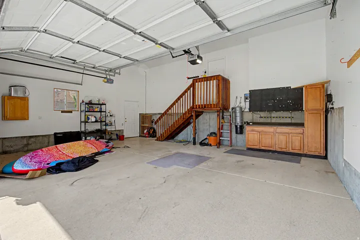 Spacious garage with polished concrete flooring, overhead sectional garage door with automatic opener, wood cabinetry storage, wooden stairs with railing providing attic access, and bright overhead lighting from recessed fixtures and skylights