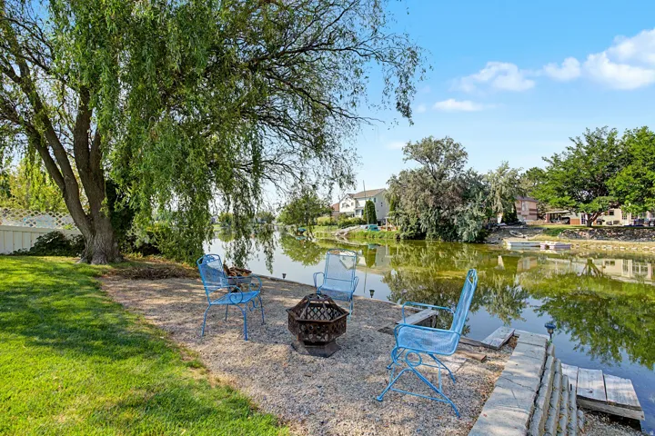 Expansive waterfront property featuring a manicured lawn, established weeping willow, gravel patio, and stone retaining wall