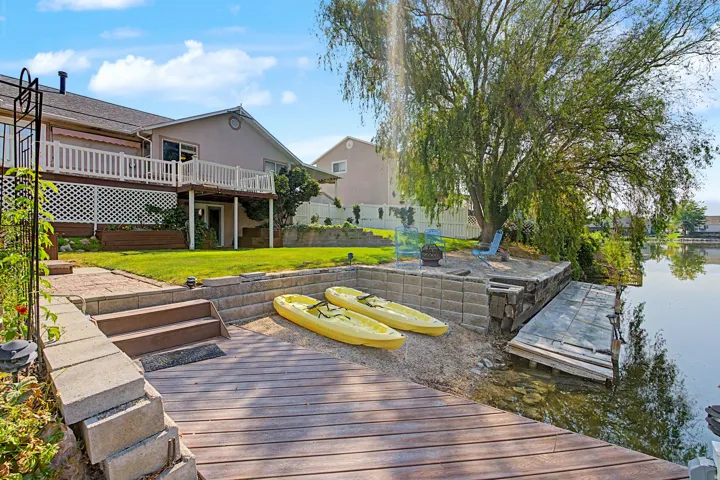 Waterside property featuring a built-in dock, slatted wood-finish decking, terraced landscaping, a mature tree, and a second-story balcony