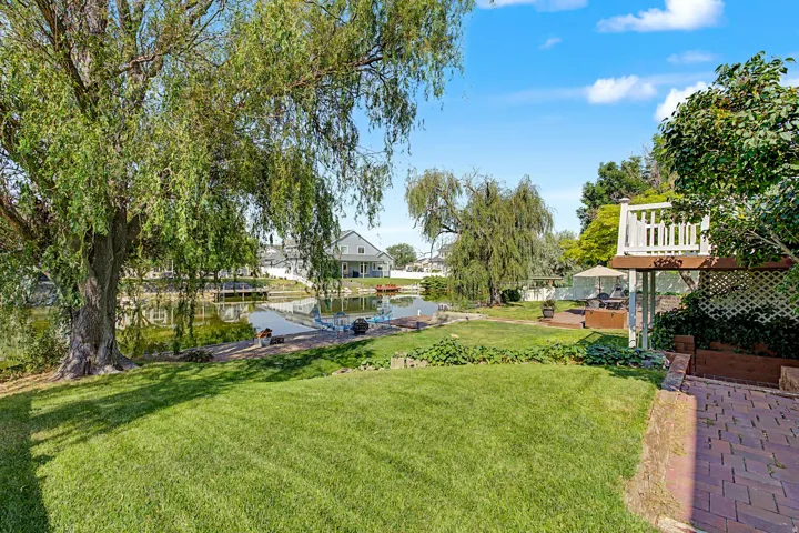 Expansive waterfront backyard featuring mature trees, a manicured lawn, a paver patio, and an elevated deck with white railings