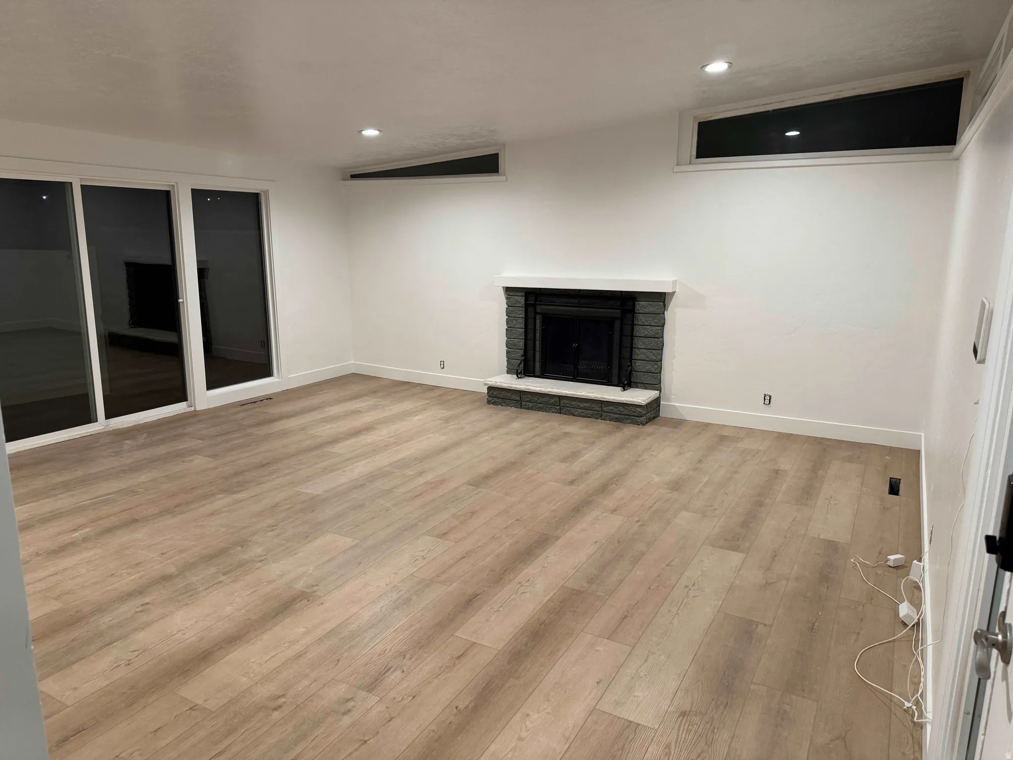 Spacious room featuring wood-finish flooring, a stone-clad fireplace with a white mantel, sliding glass doors, and recessed lighting