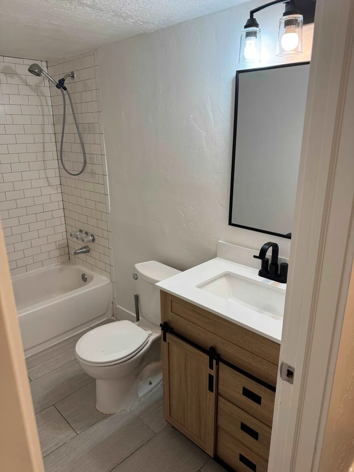Bathroom featuring a white subway tile shower surround, a built-in bathtub, a wood-finish vanity with a white countertop and integrated sink, a matte black faucet, and a black-framed mirror