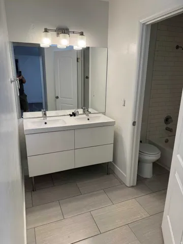 Dual vanity with integrated sinks, contemporary wall-mounted light fixture, large wall mirror, and rectangular tile flooring