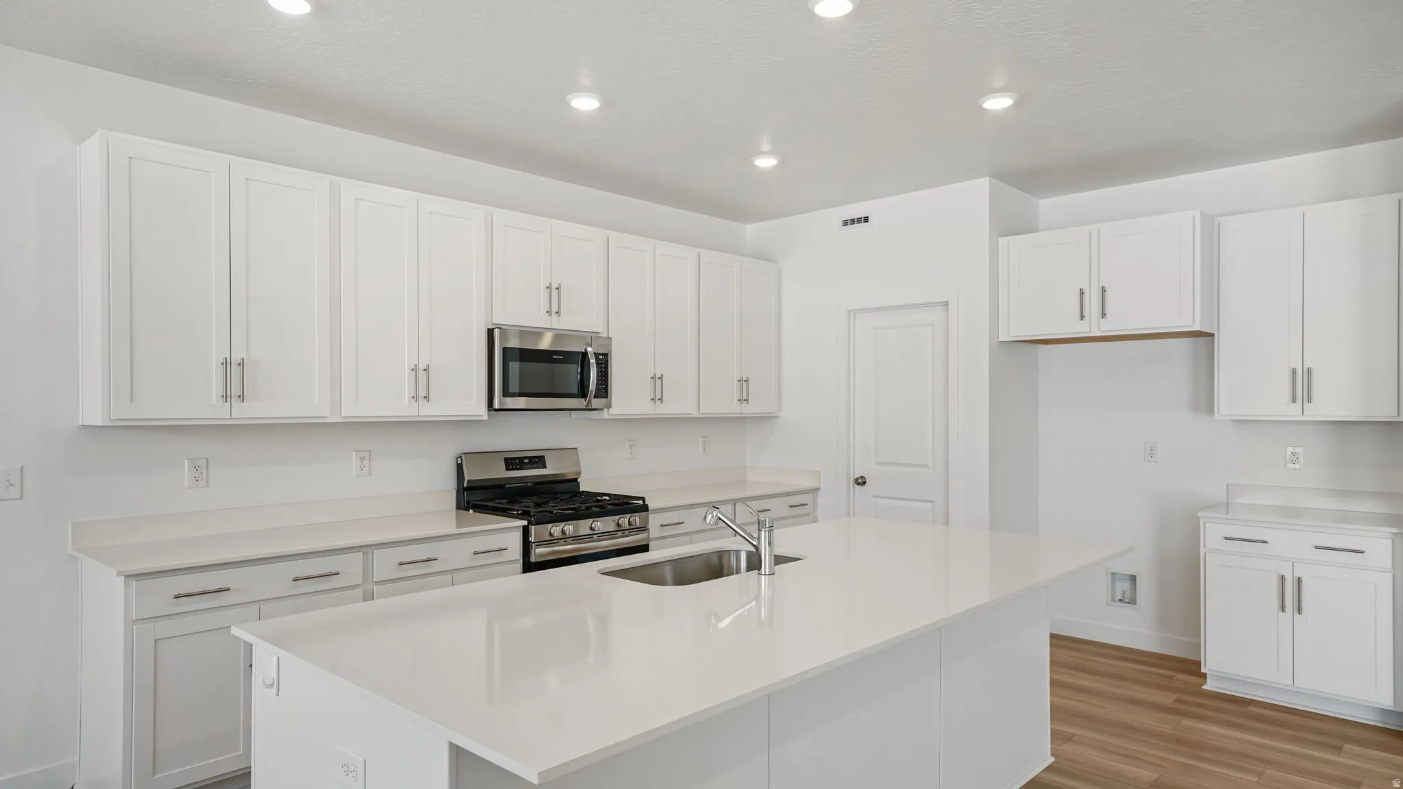 White shaker cabinetry, stainless steel appliances, central island with integrated sink, light-toned countertops, and recessed lighting