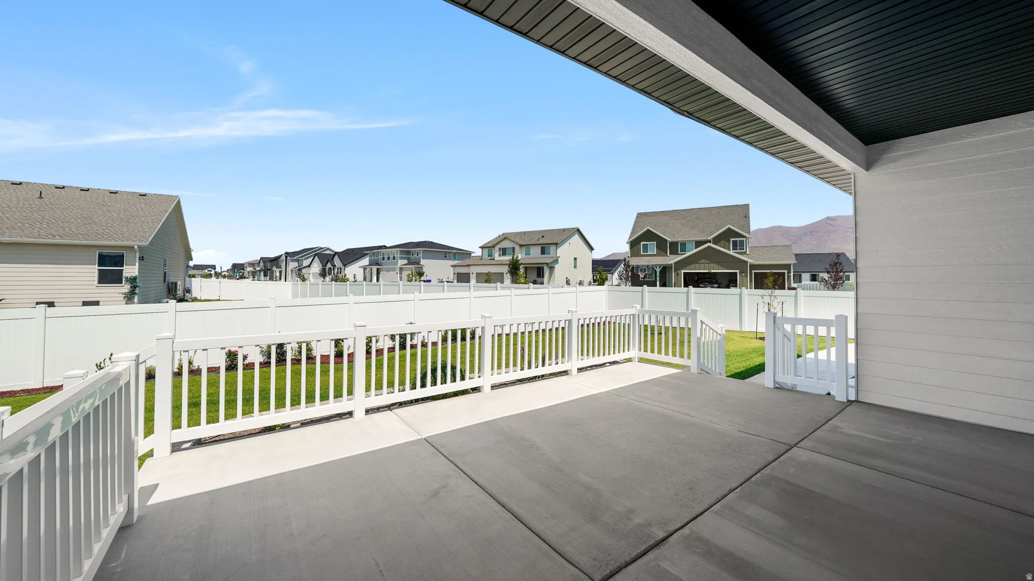 Expansive covered concrete patio with white railing and gate, featuring a manicured lawn and white privacy fencing