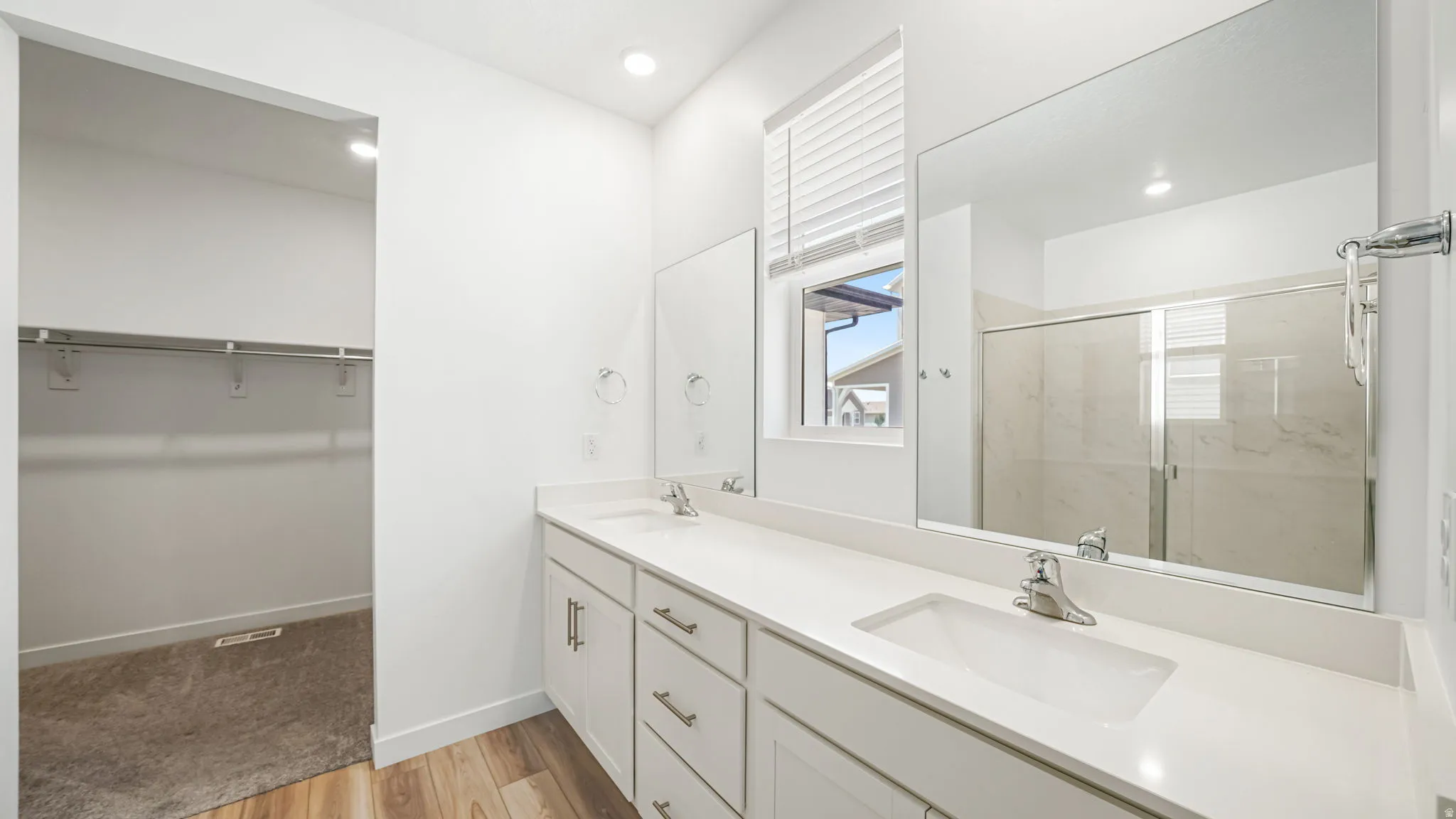 Primary suite bathroom with dual vanity with white cabinetry, wood-finish flooring, recessed lighting, and a walk-in closet and shower with sliding glass doors