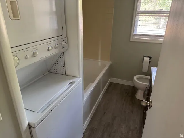 Efficient laundry and bathroom suite featuring stackable washer/dryer unit with control panel, wood-finish flooring throughout, ceramic toilet with wall-mounted paper holder, and natural light from operable window