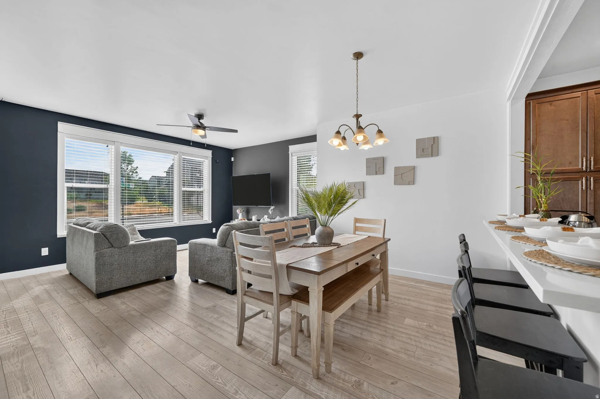 Open-concept living and dining area featuring wood-finish flooring, a dark accent wall with large multi-pane windows, a brass pendant light fixture, and direct access to adjacent kitchen cabinetry with contrasting finishes