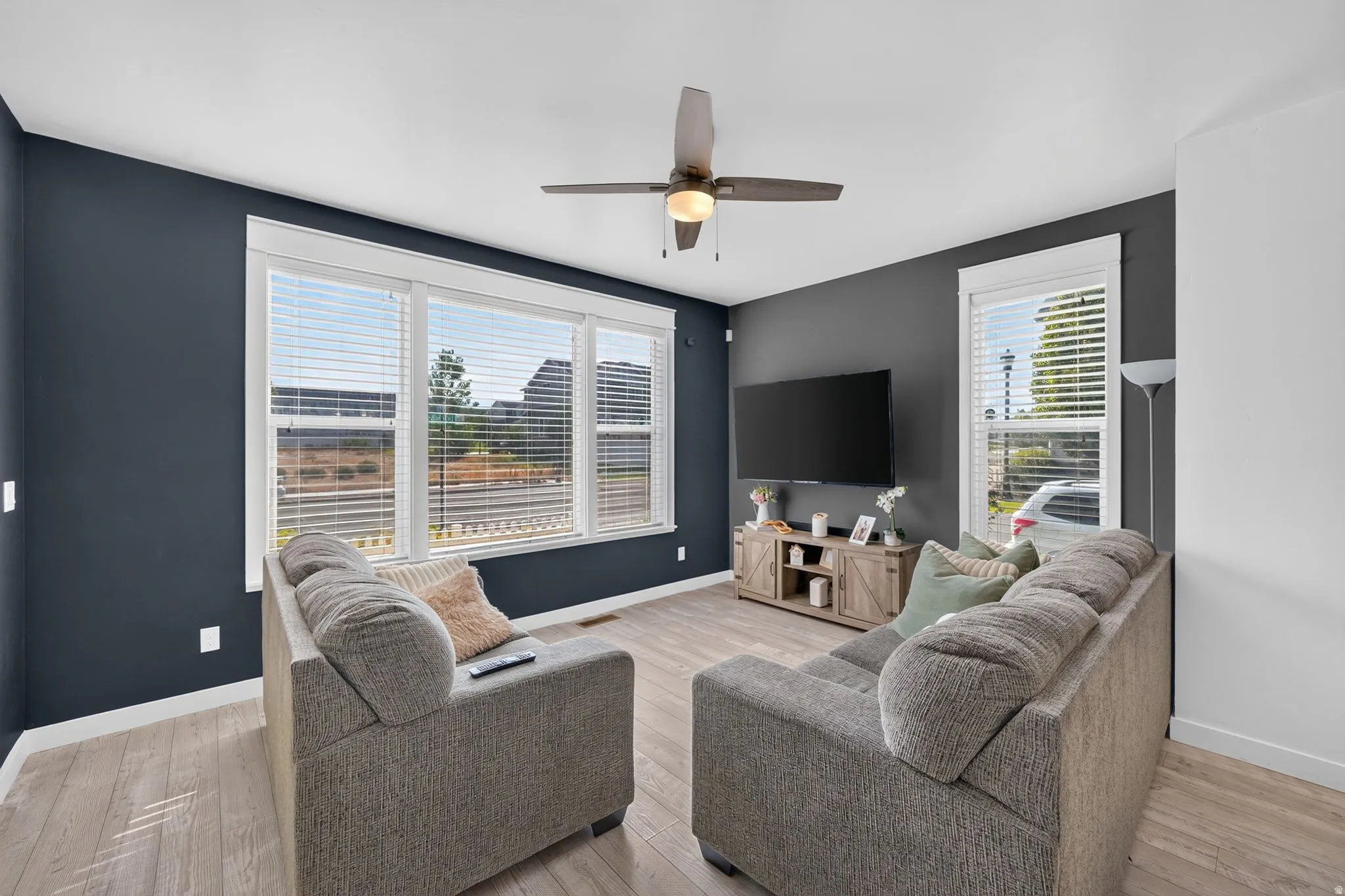 Expansive multi-pane window with white blinds, wood-finish flooring, dark accent wall, and a ceiling fan with integrated lighting
