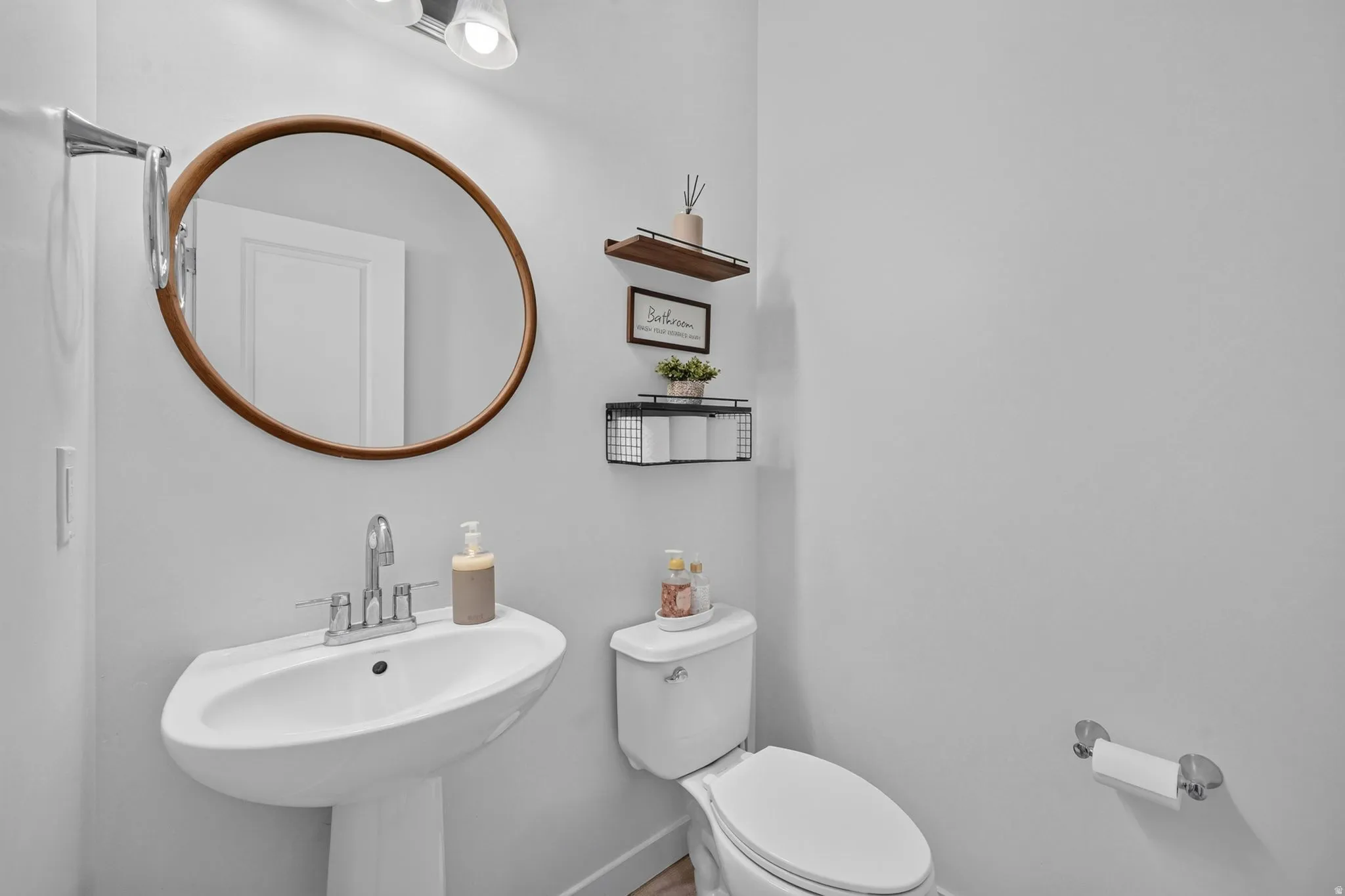 Pedestal sink with a chrome faucet, round mirror with a wood-finish frame, and built-in shelving