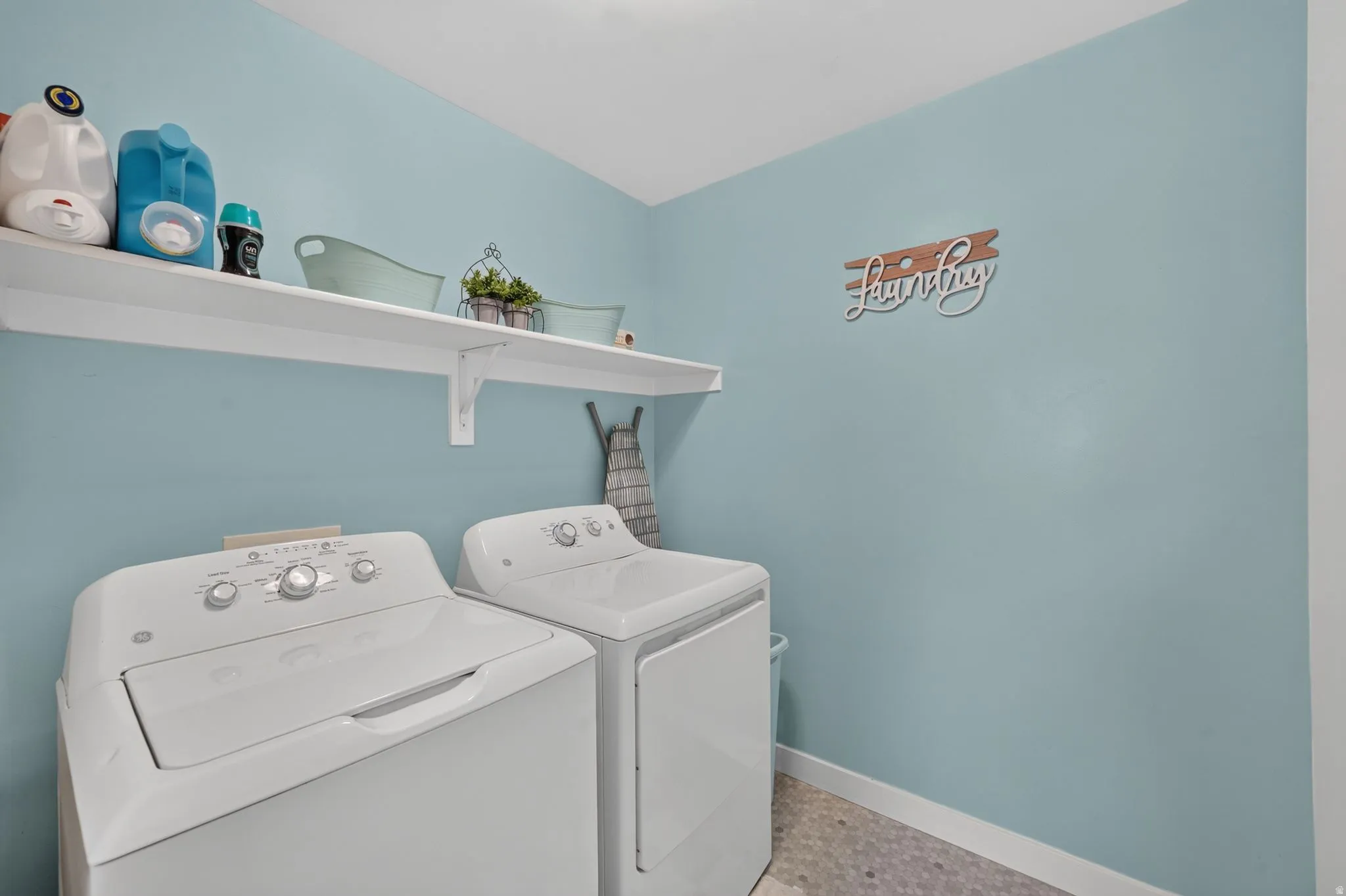 Integrated laundry appliances with upper storage shelving, light blue wall paint, and patterned floor tiling