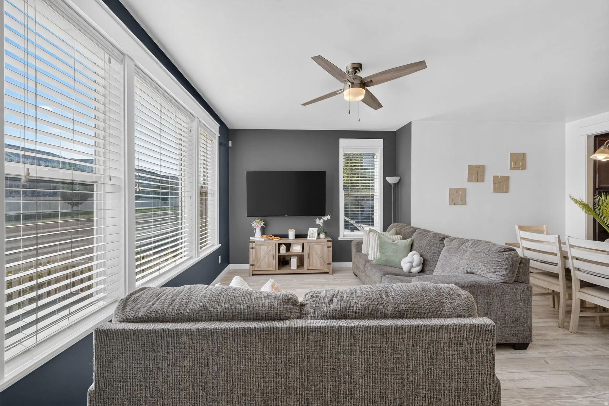 Spacious living area featuring large windows, a ceiling fan with integrated lighting, wood-finish flooring, and a media console