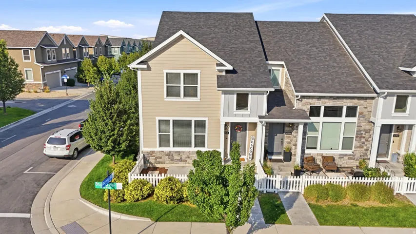 Townhome exterior featuring tan siding, a dark shingle roof, stone accenting, and a white picket fence
