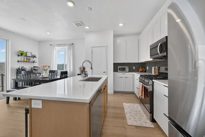 Modern kitchen featuring a light wood-finish island with a white countertop and undermount sink, white cabinetry with dark hardware, stainless steel appliances, and a dark tile backsplash