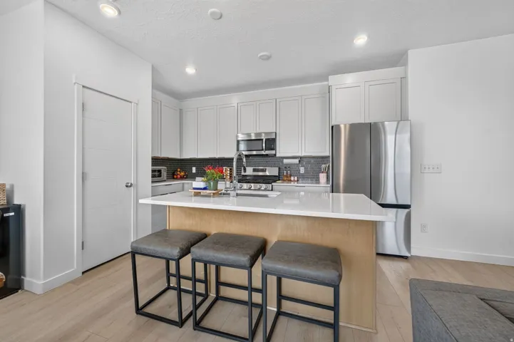 Light wood-finish flooring, white kitchen cabinetry, stainless steel appliances, white countertop, and gray tile backsplash