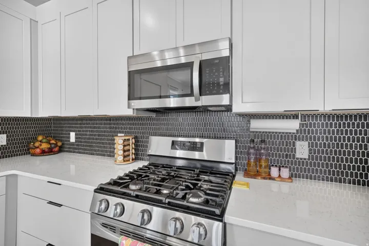Stainless steel gas range, coordinating microwave, white shaker-style cabinetry, dark geometric tile backsplash, and light-toned countertops