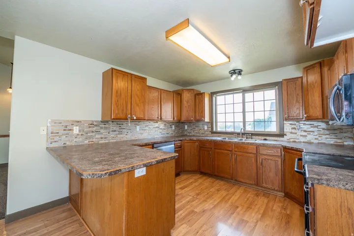 Wood-finish flooring, wood cabinetry, multi-tone tile backsplash, built-in microwave, and a window above the sink